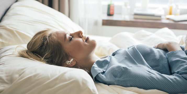 Happy blonde woman in blue pajamas lying in bed in the morning and listening to relaxing music.